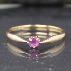 Op de afbeelding: Een gouden ring met een kleine, ovaalvormige roze edelsteen in het midden. De ring heeft een delicate band en een gepolijste afwerking, die het licht reflecteert. De edelsteen heeft een levendige roze kleur.