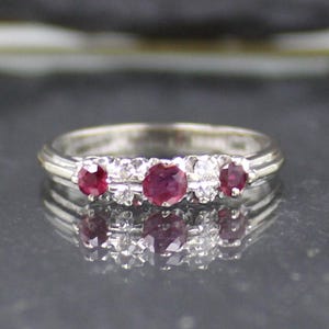 May include: A silver ring featuring three round, deep red gemstones and smaller clear stones. The ring is displayed on a reflective surface. This piece of jewellery is a ring.