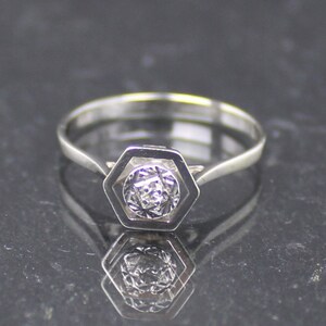 May include: A silver ring featuring a hexagonal setting. The setting contains a round, textured gemstone. The ring's band is slender and polished, reflecting light. The ring is shown on a dark, reflective surface.