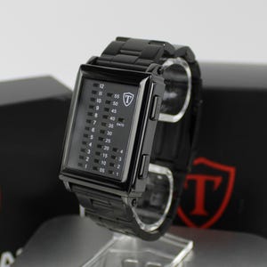 May include: A black rectangular digital watch with a metal link band. The watch face features a vertical grid display with numbers for hours, minutes, and date. A white shield logo with the letter T is visible on the upper right of the watch face.