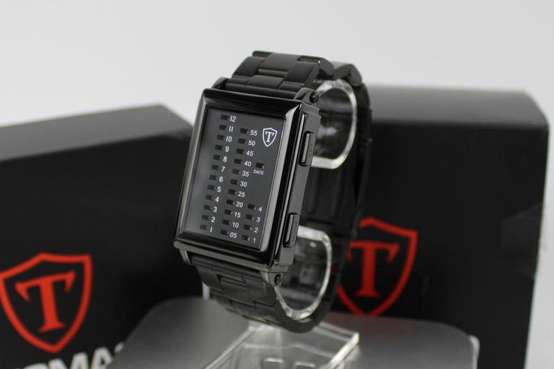 May include: A black rectangular digital watch with a metal link band. The watch face features a vertical grid display with numbers for hours, minutes, and date. A white shield logo with the letter T is visible on the upper right of the watch face.