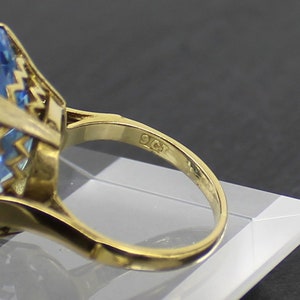 Puede incluir: Un anillo de oro con una gran piedra preciosa azul claro engastada en un engaste de oro detallado. El anillo est&aacute; marcado con "9CT" en la banda interior.