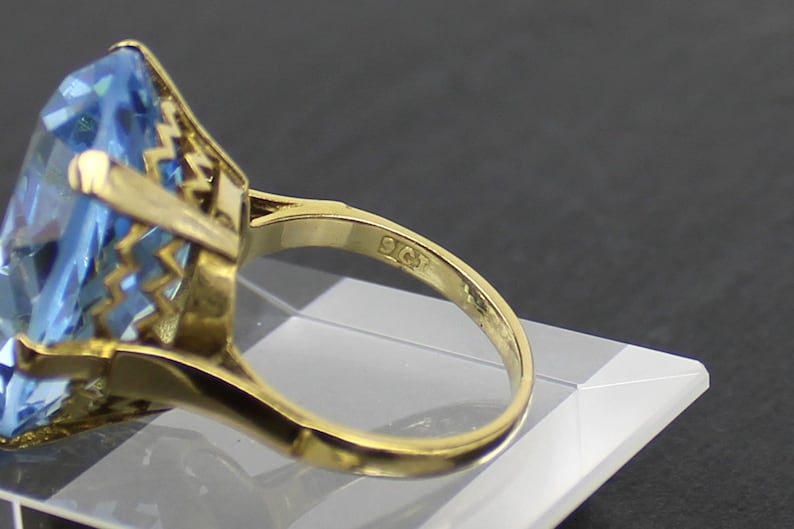 Puede incluir: Un anillo de oro con una gran piedra preciosa azul claro engastada en un engaste de oro detallado. El anillo est&aacute; marcado con "9CT" en la banda interior.