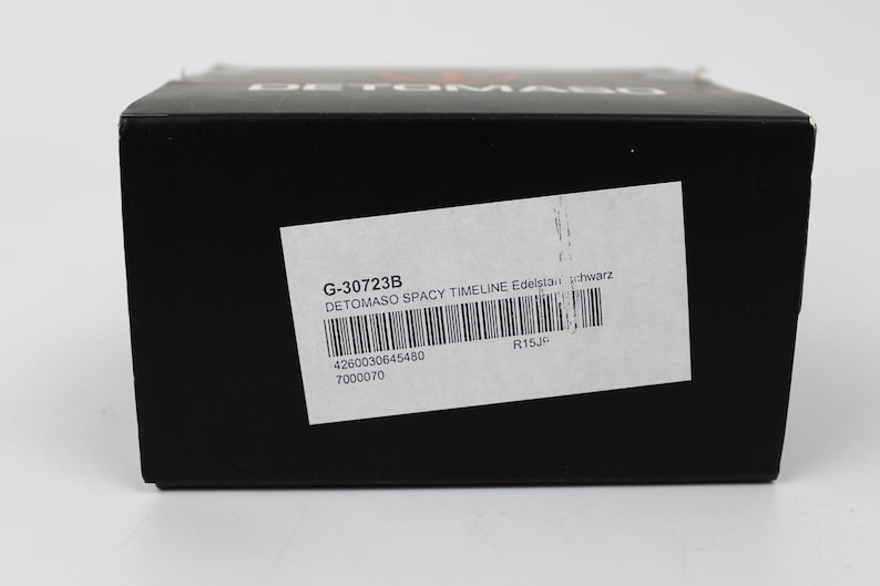 May include: A black rectangular product box with the brand name DETOMASO printed in white on the top edge. A white rectangular label is affixed to the front, featuring the text G-30723B, DETOMASO SPACY TIMELINE Edelstahl schwarz, and a barcode.
