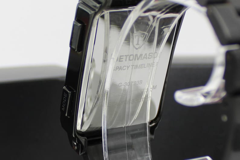 May include: A black digital watch with a rectangular case and a black strap. The back of the watch case features the text ETOMASO, LEGACY TIMELINE, G-30723B, and a shield logo. The watch is mounted on a clear plastic display stand.