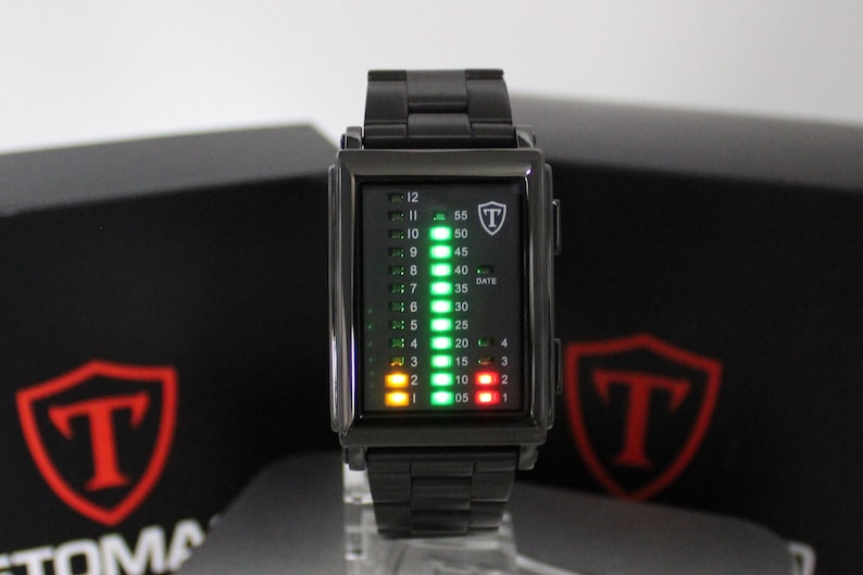 May include: A black rectangular digital watch with a metal link band. The watch face features a vertical LED display with green, yellow, and red lights indicating time and date. A shield logo with the letter T is visible on the upper right corner.