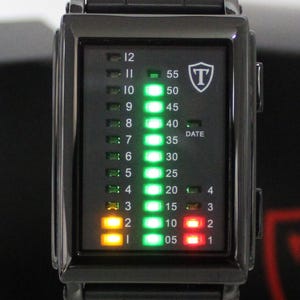 May include: A rectangular black digital watch with a vertical LED display. The face features columns of numbers for hours 1 through 12, minutes 05 through 55, and a date indicator. Bright green, yellow, and red lights illuminate the display, with a shield logo containing the letter T in the upper right corner.