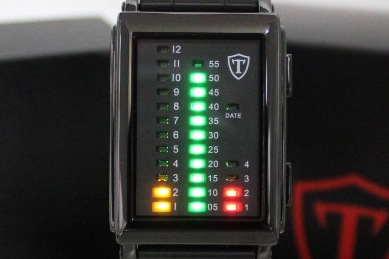 May include: A rectangular black digital watch with a vertical LED display. The face features columns of numbers for hours 1 through 12, minutes 05 through 55, and a date indicator. Bright green, yellow, and red lights illuminate the display, with a shield logo containing the letter T in the upper right corner.