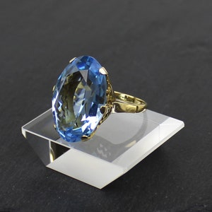 Puede incluir: Un anillo de oro con una gran piedra preciosa ovalada de color azul claro. El anillo est&aacute; en un expositor blanco.
