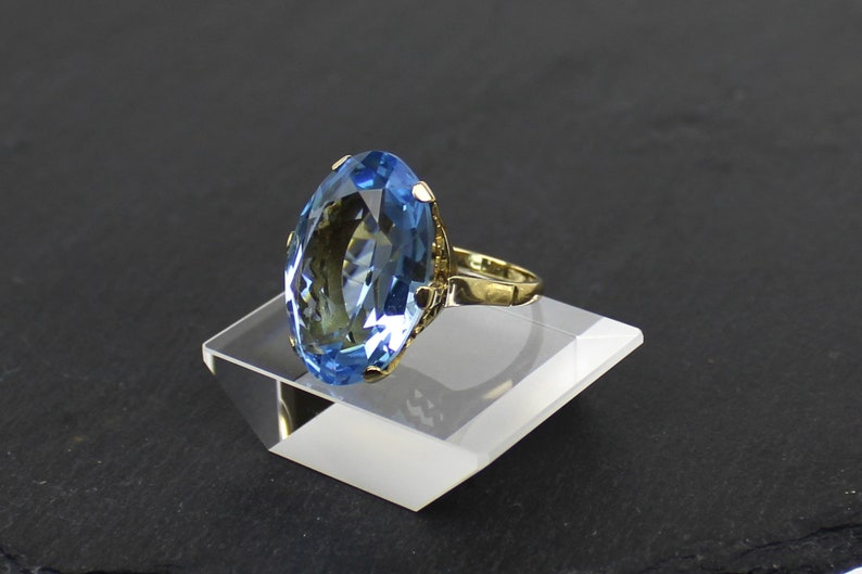Puede incluir: Un anillo de oro con una gran piedra preciosa ovalada de color azul claro. El anillo est&aacute; en un expositor blanco.