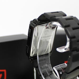 May include: A black wristwatch with a rectangular case and a matching black link bracelet. The back of the watch case is metallic and features the engraved text "TOMASO" along with a shield logo. The watch is displayed on a clear stand.