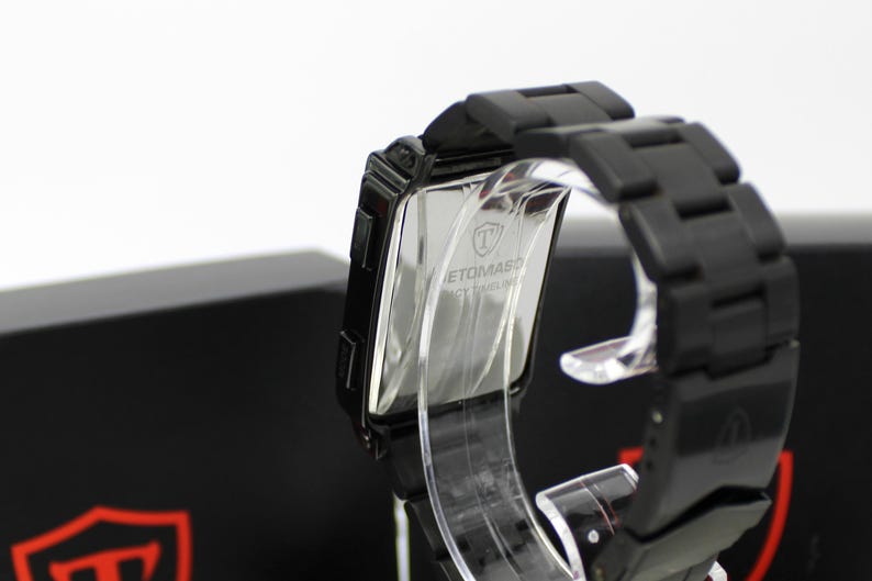 May include: A black wristwatch with a rectangular case and a matching black link bracelet. The back of the watch case is metallic and features the engraved text "TOMASO" along with a shield logo. The watch is displayed on a clear stand.