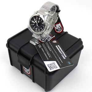 K&ouml;nnte beinhalten: Eine silberne Uhr mit schwarzem Zifferblatt und einem Mesh-Armband. Die Uhr befindet sich in einem schwarzen Etui mit dem Logo "LUMINOX". Das Etui hat eine Garantiekarte mit der Aufschrift "INTERNATIONAL WARRANTY CARD" befestigt.