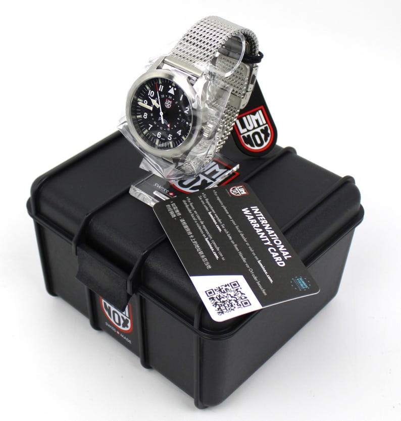 K&ouml;nnte beinhalten: Eine silberne Uhr mit schwarzem Zifferblatt und einem Mesh-Armband. Die Uhr befindet sich in einem schwarzen Etui mit dem Logo "LUMINOX". Das Etui hat eine Garantiekarte mit der Aufschrift "INTERNATIONAL WARRANTY CARD" befestigt.