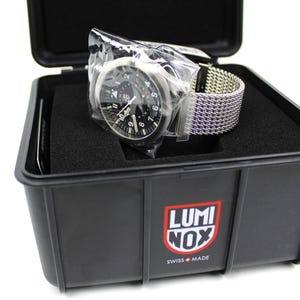 K&ouml;nnte beinhalten: Eine schwarze Luminox-Uhr mit schwarzem Zifferblatt und silbernen Zeigern und Markierungen. Die Uhr befindet sich in einem schwarzen Kunststoffgeh&auml;use mit dem Luminox-Logo auf der Vorderseite. Die Uhr hat ein silbernes Mesh-Armband.