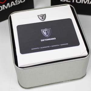 May include: A square silver metal storage tin containing a white instruction booklet and a black warranty card. The card features the Detomaso logo, a shield with the letter T, and the text GARANTE, GUARANTEE, GARANTIA, GARANZIA printed below.