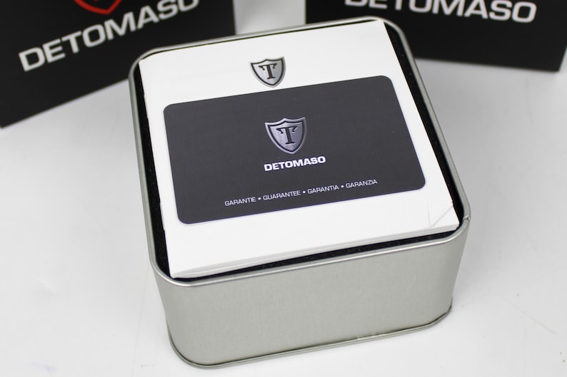 May include: A square silver metal storage tin containing a white instruction booklet and a black warranty card. The card features the Detomaso logo, a shield with the letter T, and the text GARANTE, GUARANTEE, GARANTIA, GARANZIA printed below.