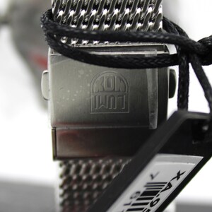 K&ouml;nnte beinhalten: Nahaufnahme einer silbernen Uhr mit einem schwarzen Mesh-Armband. Das Zifferblatt der Uhr ist nicht sichtbar, aber auf der R&uuml;ckseite des Uhrengeh&auml;uses sind die Worte "KOM  WUT" eingraviert. Die Uhr ist an einem schwarzen Etikett mit einem Barcode befestigt.