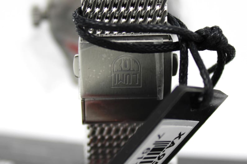 K&ouml;nnte beinhalten: Nahaufnahme einer silbernen Uhr mit einem schwarzen Mesh-Armband. Das Zifferblatt der Uhr ist nicht sichtbar, aber auf der R&uuml;ckseite des Uhrengeh&auml;uses sind die Worte "KOM  WUT" eingraviert. Die Uhr ist an einem schwarzen Etikett mit einem Barcode befestigt.