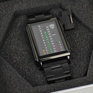 May include: A black rectangular digital watch with a metal link band, featuring a vertical LED display with green and orange light indicators for hours and minutes, and a shield logo with the letter T at the top, presented in a foam-lined case.