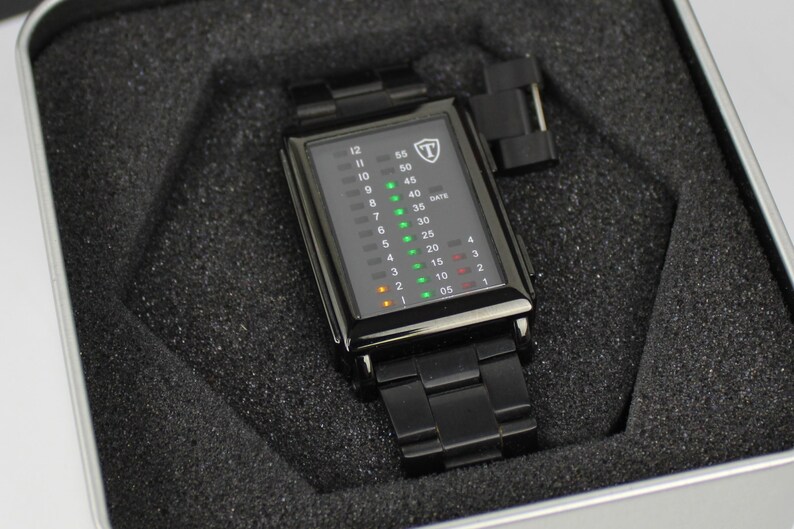 May include: A black rectangular digital watch with a metal link band, featuring a vertical LED display with green and orange light indicators for hours and minutes, and a shield logo with the letter T at the top, presented in a foam-lined case.