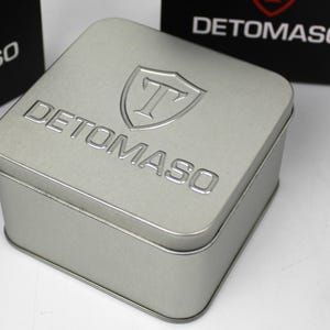 May include: A square silver-colored metal storage tin with rounded corners. The lid features the embossed text DETOMASO and a shield logo with the letter T inside. Two black boxes with the same white logo and text are visible in the background.