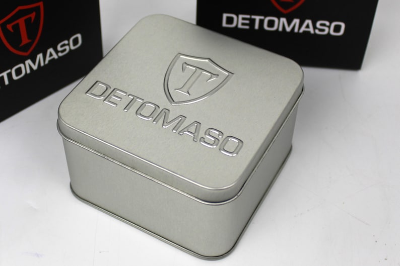 May include: A square silver-colored metal storage tin with rounded corners. The lid features the embossed text DETOMASO and a shield logo with the letter T inside. Two black boxes with the same white logo and text are visible in the background.