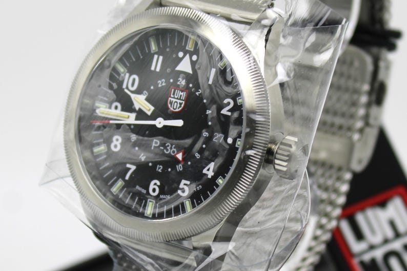 K&ouml;nnte beinhalten: Eine silberne Armbanduhr mit schwarzem Zifferblatt und wei&szlig;en Markierungen. Die Uhr hat ein schwarzes Lederarmband und eine silberne Schlie&szlig;e. Auf dem Zifferblatt der Uhr sind die Worte "LUMI NOX" und "P-36" aufgedruckt.