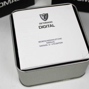 May include: A square silver-colored metal tin containing a white instruction manual. The manual features a shield logo with the letter T, the brand name DETOMASO, the word DIGITAL, and the text BEDIENUNGSANLEITUNG, MANUAL, MANUEL D'UTILISATION.