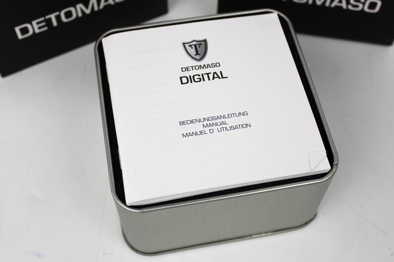 May include: A square silver-colored metal tin containing a white instruction manual. The manual features a shield logo with the letter T, the brand name DETOMASO, the word DIGITAL, and the text BEDIENUNGSANLEITUNG, MANUAL, MANUEL D'UTILISATION.