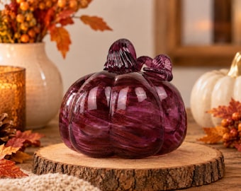 Könnte beinhalten: Eine dunkelviolette Glaskürbis-Skulptur mit gerippter Oberfläche und strukturiertem Stiel. Das Glas weist wirbelnde dunkle und helle Violetttöne auf, die einen durchscheinenden Effekt erzeugen. Der Kürbis steht auf einem runden Holzsockel.