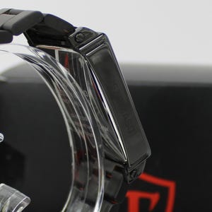 May include: A matte black wristwatch featuring a rectangular case and a matching link bracelet. The side of the watch case displays the brand name DETOMASO engraved in a vertical orientation. The watch is mounted on a clear display stand.