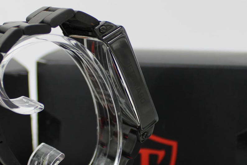 May include: A matte black wristwatch featuring a rectangular case and a matching link bracelet. The side of the watch case displays the brand name DETOMASO engraved in a vertical orientation. The watch is mounted on a clear display stand.