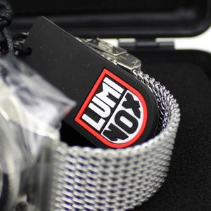 K&ouml;nnte beinhalten: Ein silbernes Metallarmband mit einem schwarzen Zifferblatt und einem schwarzen Etikett mit dem Wort "LUMINOX" in roten Buchstaben.
