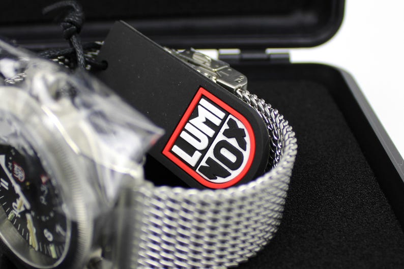 K&ouml;nnte beinhalten: Ein silbernes Metallarmband mit einem schwarzen Zifferblatt und einem schwarzen Etikett mit dem Wort "LUMINOX" in roten Buchstaben.