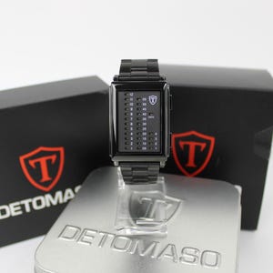 May include: A black rectangular digital watch with a metal link band. The watch face features a vertical binary-style LED display with numbers 1 through 12 on the left and increments of 5 on the right, marked with the Detomaso logo and text.