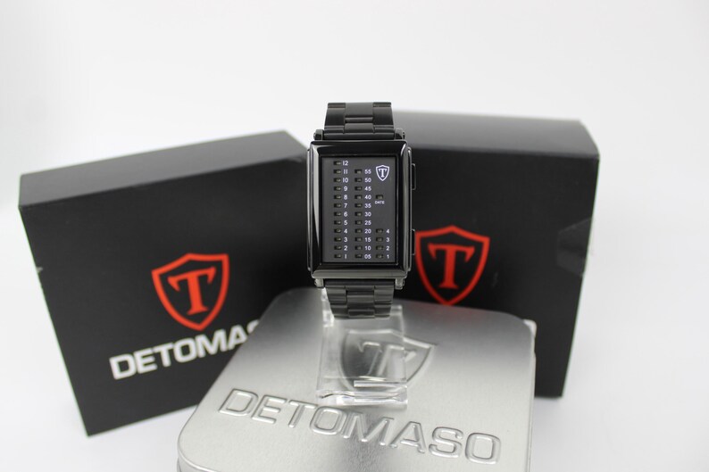 May include: A black rectangular digital watch with a metal link band. The watch face features a vertical binary-style LED display with numbers 1 through 12 on the left and increments of 5 on the right, marked with the Detomaso logo and text.