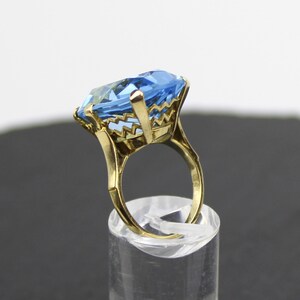 Puede incluir: Un anillo de oro con una gran piedra preciosa azul claro facetada. El anillo tiene un dise&ntilde;o delicado e intrincado alrededor de la piedra.