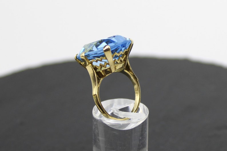Puede incluir: Un anillo de oro con una gran piedra preciosa azul claro facetada. El anillo tiene un dise&ntilde;o delicado e intrincado alrededor de la piedra.