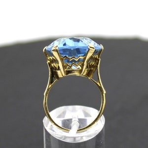 Puede incluir: Un anillo de oro con una gran piedra preciosa azul claro facetada. El anillo est&aacute; colocado en un expositor transparente.