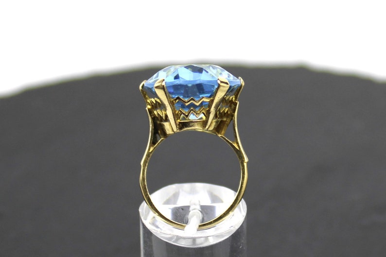 Puede incluir: Un anillo de oro con una gran piedra preciosa azul claro facetada. El anillo est&aacute; colocado en un expositor transparente.