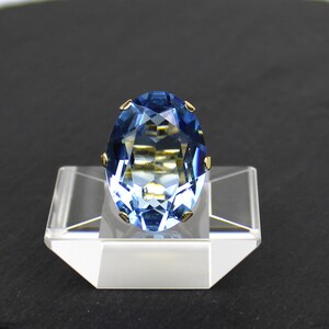 Puede incluir: Un anillo de piedra ovalada azul con detalles dorados. El anillo est&aacute; colocado en un expositor transparente.