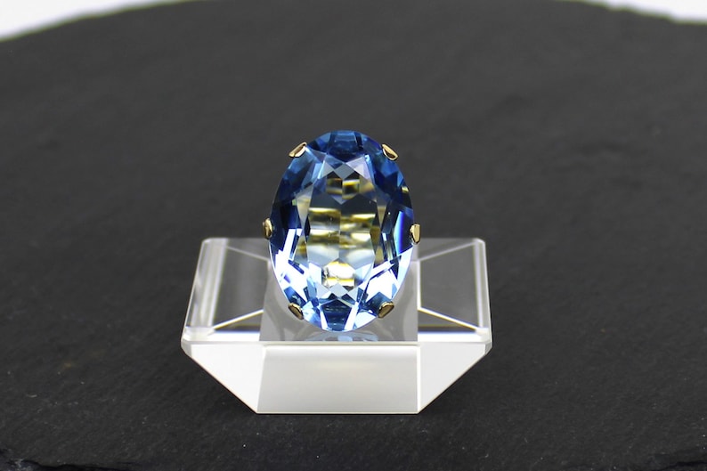 Puede incluir: Un anillo de piedra ovalada azul con detalles dorados. El anillo est&aacute; colocado en un expositor transparente.