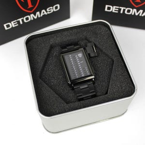 May include: A black rectangular Detomaso wristwatch with a digital-style vertical display featuring numbers and a shield logo on the dial. The watch has a matching black metal link band and is presented inside a square silver metal tin with foam.