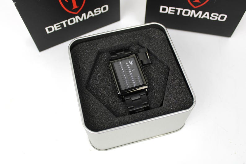 May include: A black rectangular Detomaso wristwatch with a digital-style vertical display featuring numbers and a shield logo on the dial. The watch has a matching black metal link band and is presented inside a square silver metal tin with foam.