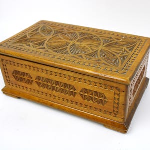 Könnte beinhalten: Eine rechteckige Holzbox mit aufwendig geschnitzten geometrischen Mustern. Die Box hat eine warme, mittelbraune Farbe und weist detaillierte Designs auf dem Deckel und den Seiten auf. Die Box hat einen Klappdeckel und einen leicht erhöhten Sockel.