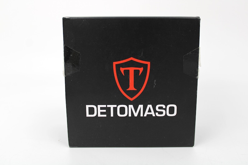 May include: A square black box featuring the white text DETOMASO centered at the bottom. Above the text is a red shield emblem containing a large red letter T. The box has a smooth, matte finish and appears to be a product packaging container.