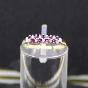 Könnte beinhalten: Ein goldener Ring mit einer Reihe kleiner, leuchtend roter Edelsteine, die mit kleineren, klaren Steinen durchsetzt sind. Der Ring wird auf einem transparenten, zylindrischen Ständer vor dunklem Hintergrund präsentiert.