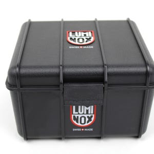 K&ouml;nnte beinhalten: Eine schwarze Plastik-Uhrenbox mit dem Luminox-Logo oben und unten. Das Logo ist rot und wei&szlig; mit dem Text "Luminox" und "Swiss Made".
