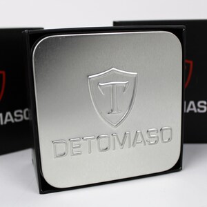 May include: A square silver metal tin featuring an embossed shield logo with the letter T and the text DETOMASO. The tin is positioned in front of two black boxes, each displaying a red shield logo with a T and the white text DETOMASO.